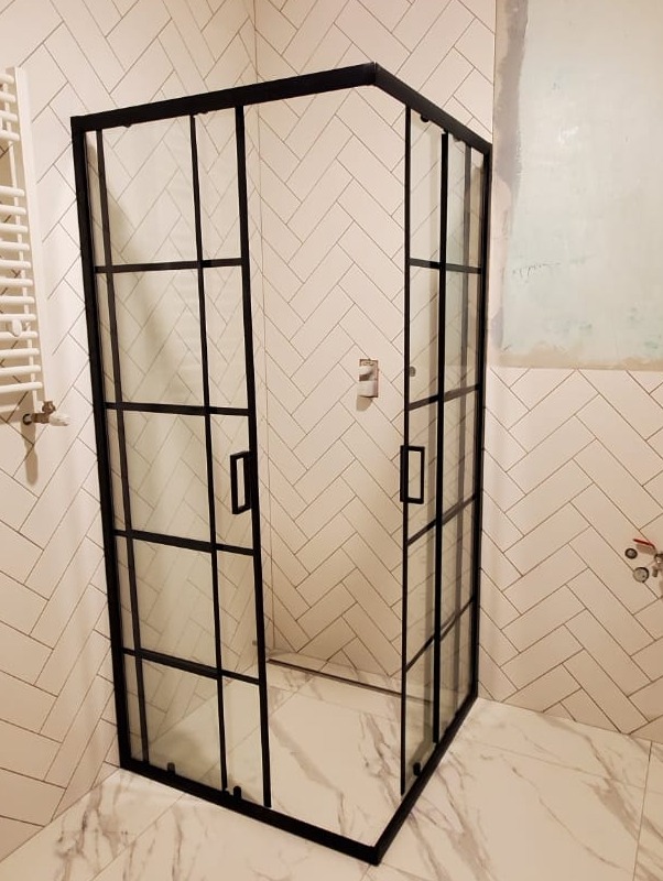 Nowoczesna kabina prysznicowa z czarnymi profilami i szklanymi ściankami, na tle białych płytek ułożonych w jodełkę i marmurowej podłogi, z widocznym grzejnikiem drabinkowym.