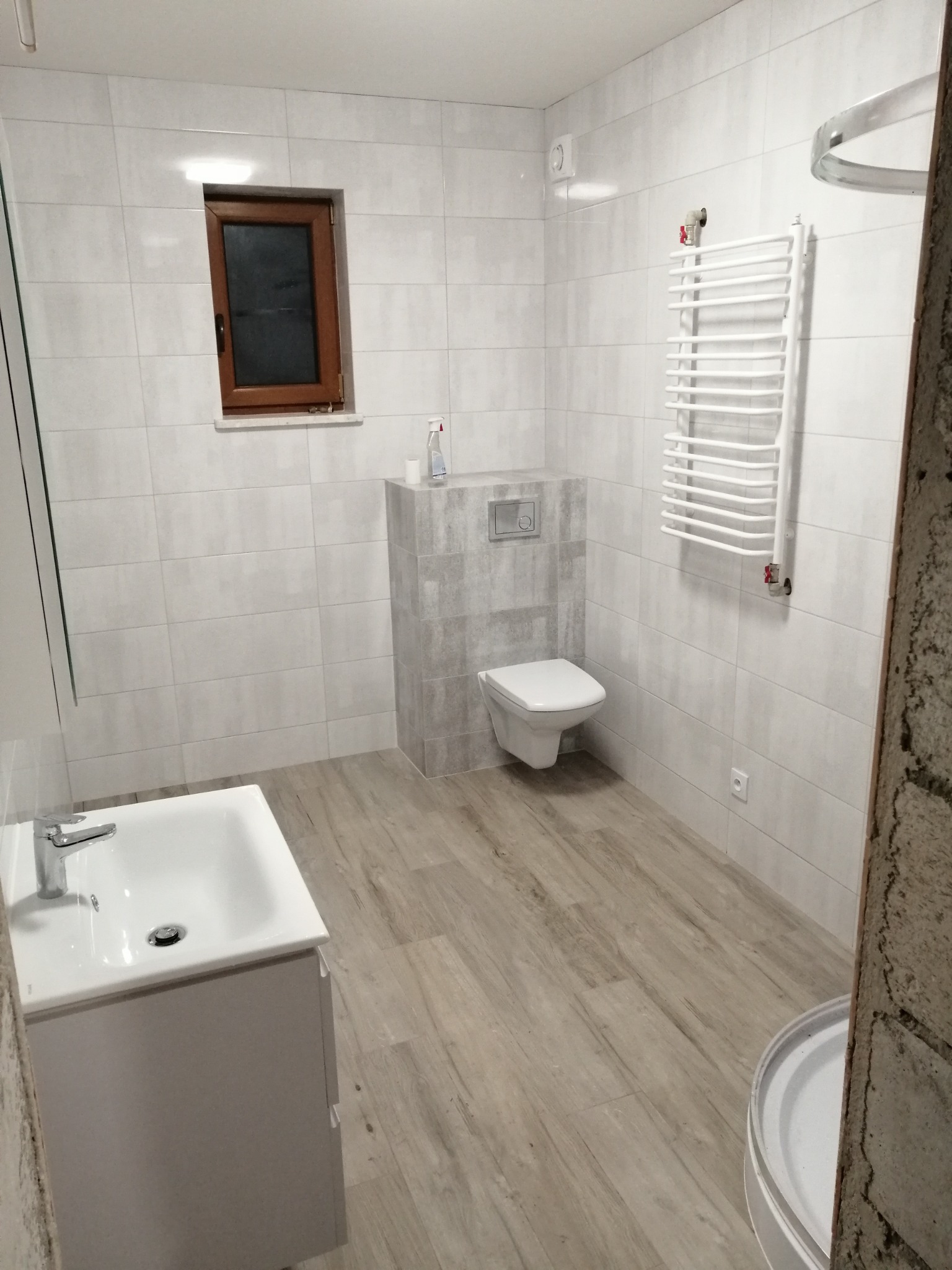 Jasna łazienka z białymi płytkami i drewnopodobną podłogą, widoczna umywalka z szafką, toaleta podtynkowa i grzejnik drabinkowy, małe okno z brązową ramą, ściany wykończone płytkami w odcieniach...