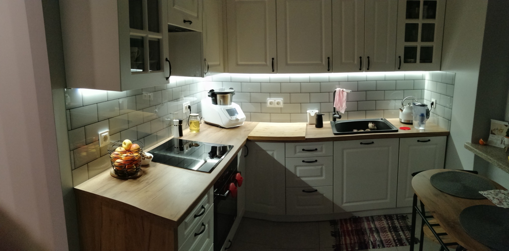 Jasna kuchnia z białymi szafkami, drewnianymi blatami i podświetleniem LED pod szafkami wiszącymi, widoczne płytki ceramiczne na ścianie, koszyk z owocami oraz sprzęt AGD na blacie.