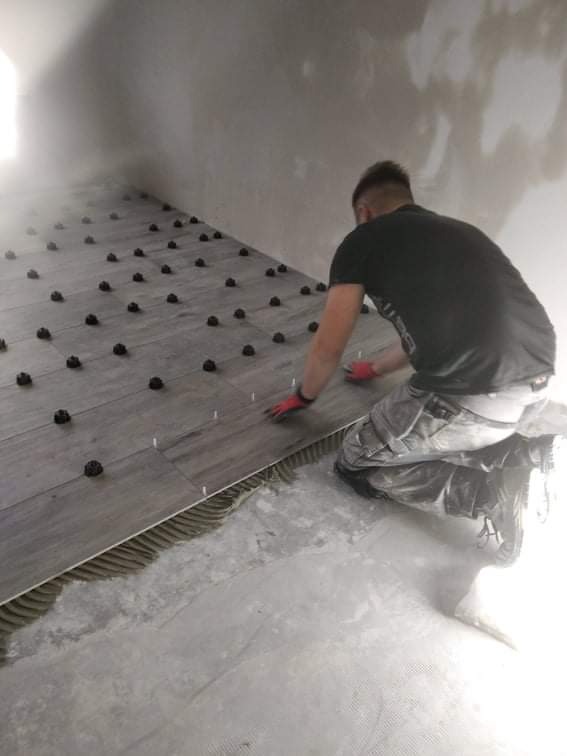 Glazurnik w trakcie układania szarych płytek podłogowych z użyciem systemu poziomowania, widoczne krzyżyki dystansowe i czarne klipsy.