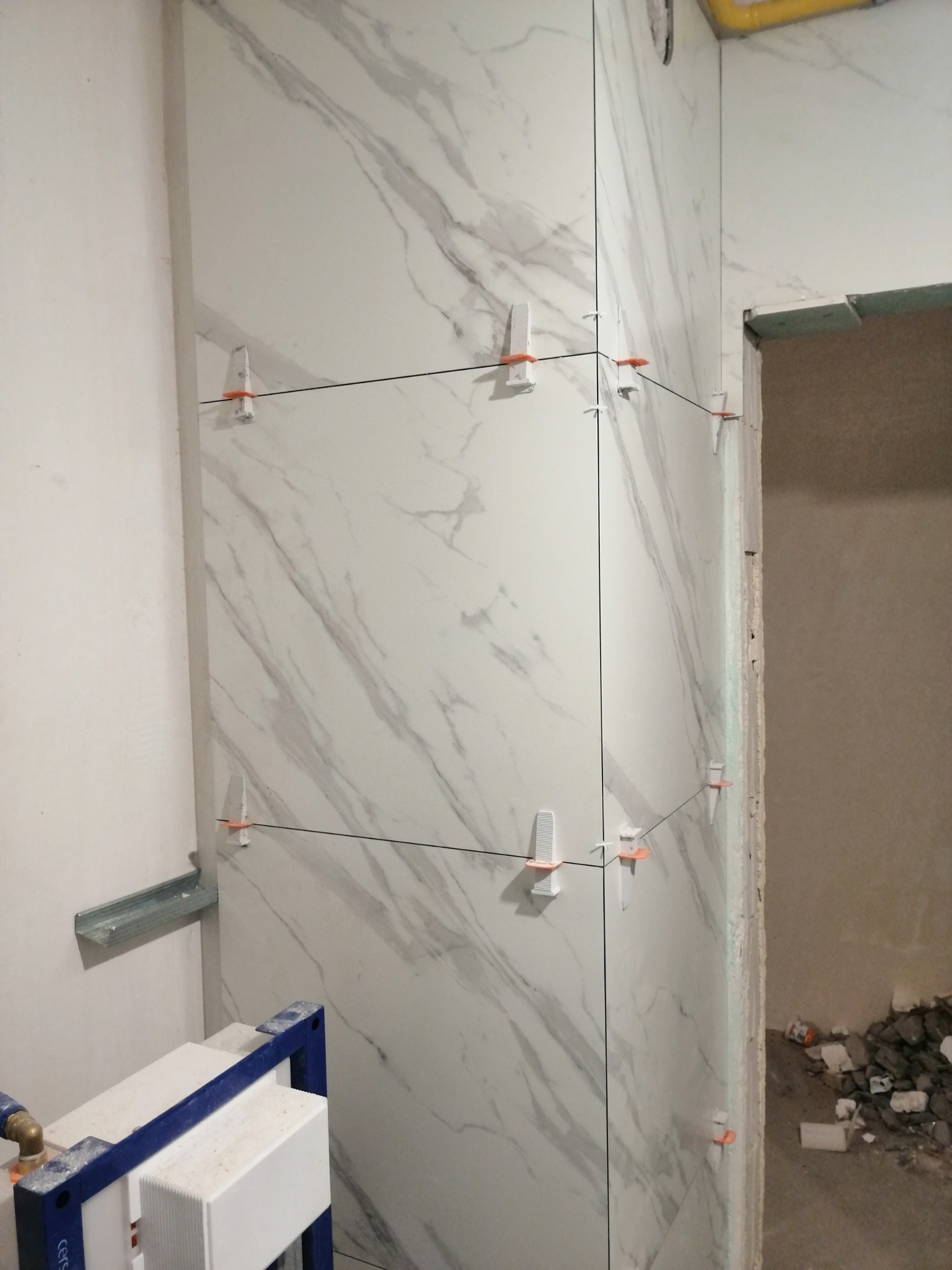 Ściana w trakcie wykańczania płytkami imitującymi marmur, z użyciem klipsów poziomujących, widoczne elementy instalacji sanitarnej.