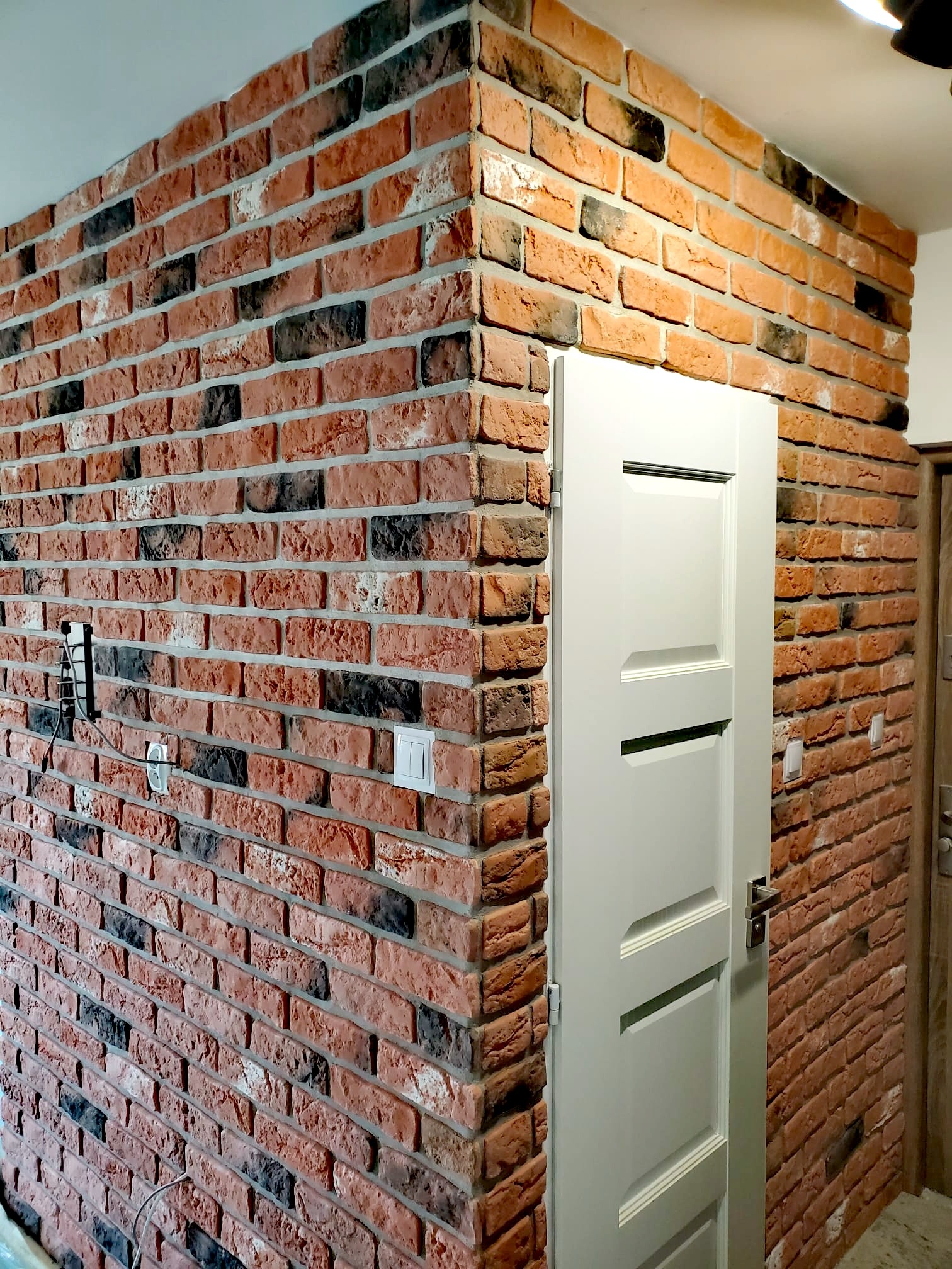 Ściana wykończona cegłą dekoracyjną w odcieniach czerwieni i czerni, widoczny fragment białych drzwi z trzema poziomymi wgłębieniami, gniazdko elektryczne i włącznik światła na ceglanej ścianie.
