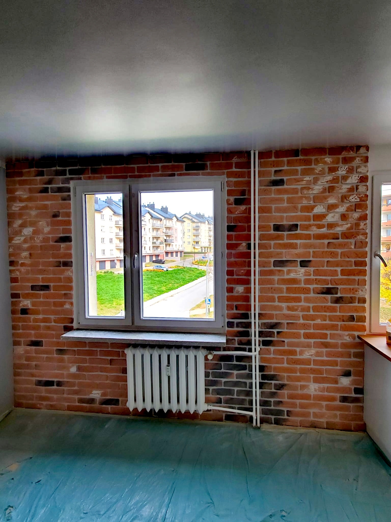 Ściana wykończona czerwoną cegłą klinkierową z ciemnymi przebarwieniami, okno z widokiem na osiedle, grzejnik pod oknem, podłoga zabezpieczona folią malarską.