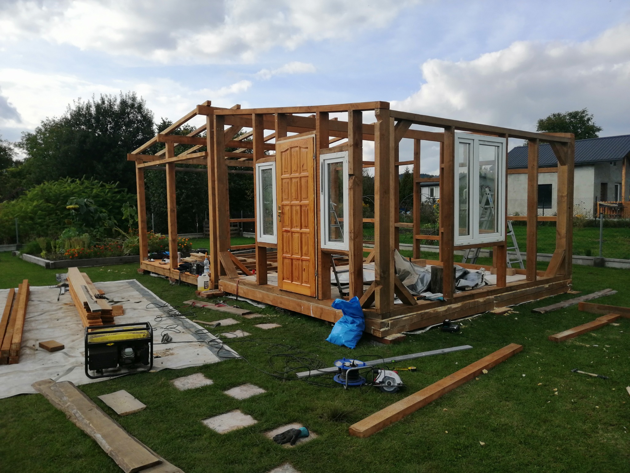 Drewniana konstrukcja altany w trakcie budowy, widoczne ramy okienne, drzwi, generator prądu, narzędzia na trawie