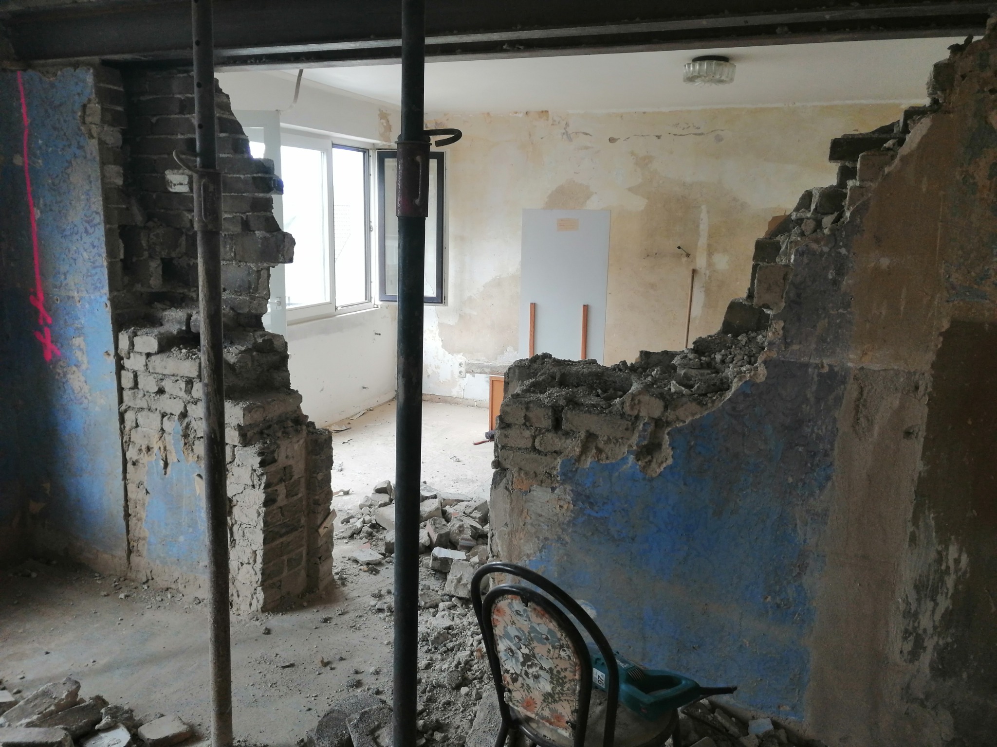 Wnętrze w trakcie demontażu ściany z cegły, widoczne podparcie stropu stalowymi stemplami, gruz na podłodze, fragment krzesła z kwiatowym wzorem i narzędzie elektryczne w tle.