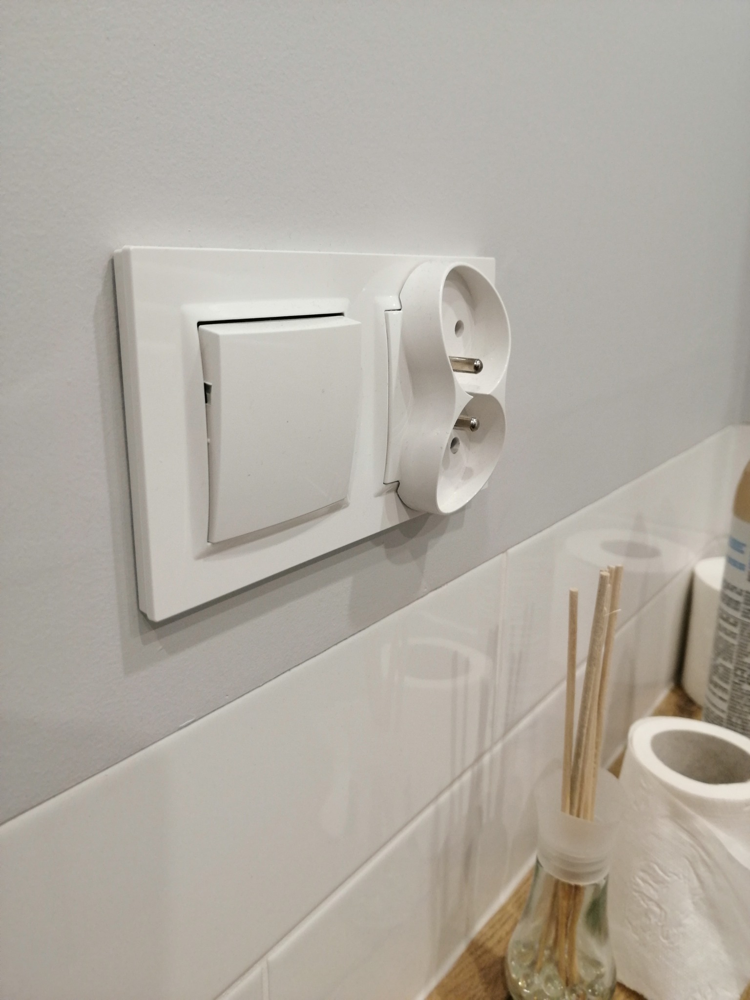 Biały włącznik światła i podwójne gniazdko elektryczne zamontowane na szarej ścianie nad rzędem białych płytek, obok flakon z patyczkami zapachowymi i rolka papieru toaletowego.