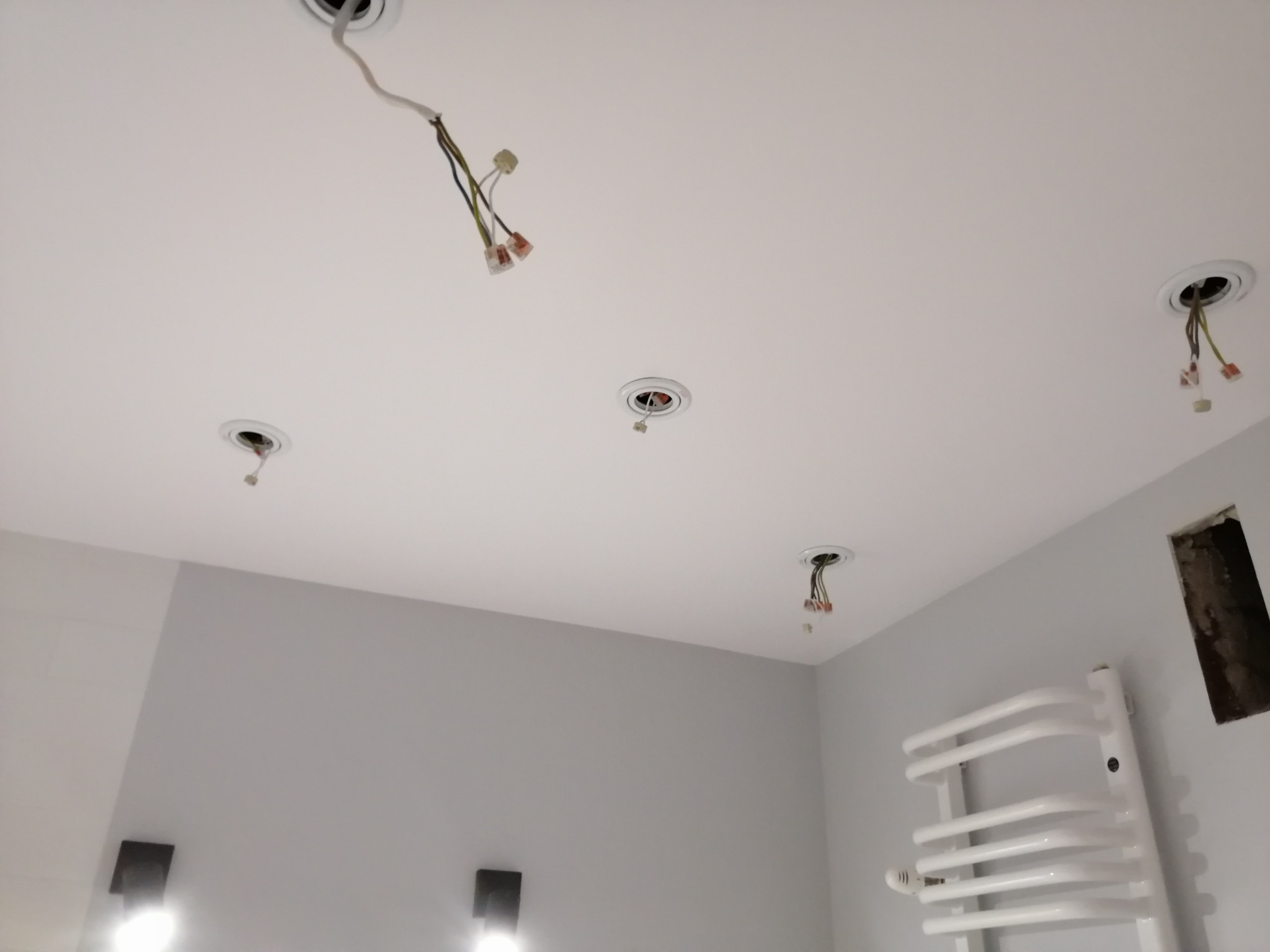 Biały sufit z widocznymi otworami na oświetlenie, z których wystają przewody z kostkami elektrycznymi, na szarej ścianie widoczny grzejnik drabinkowy i otwór w ścianie.