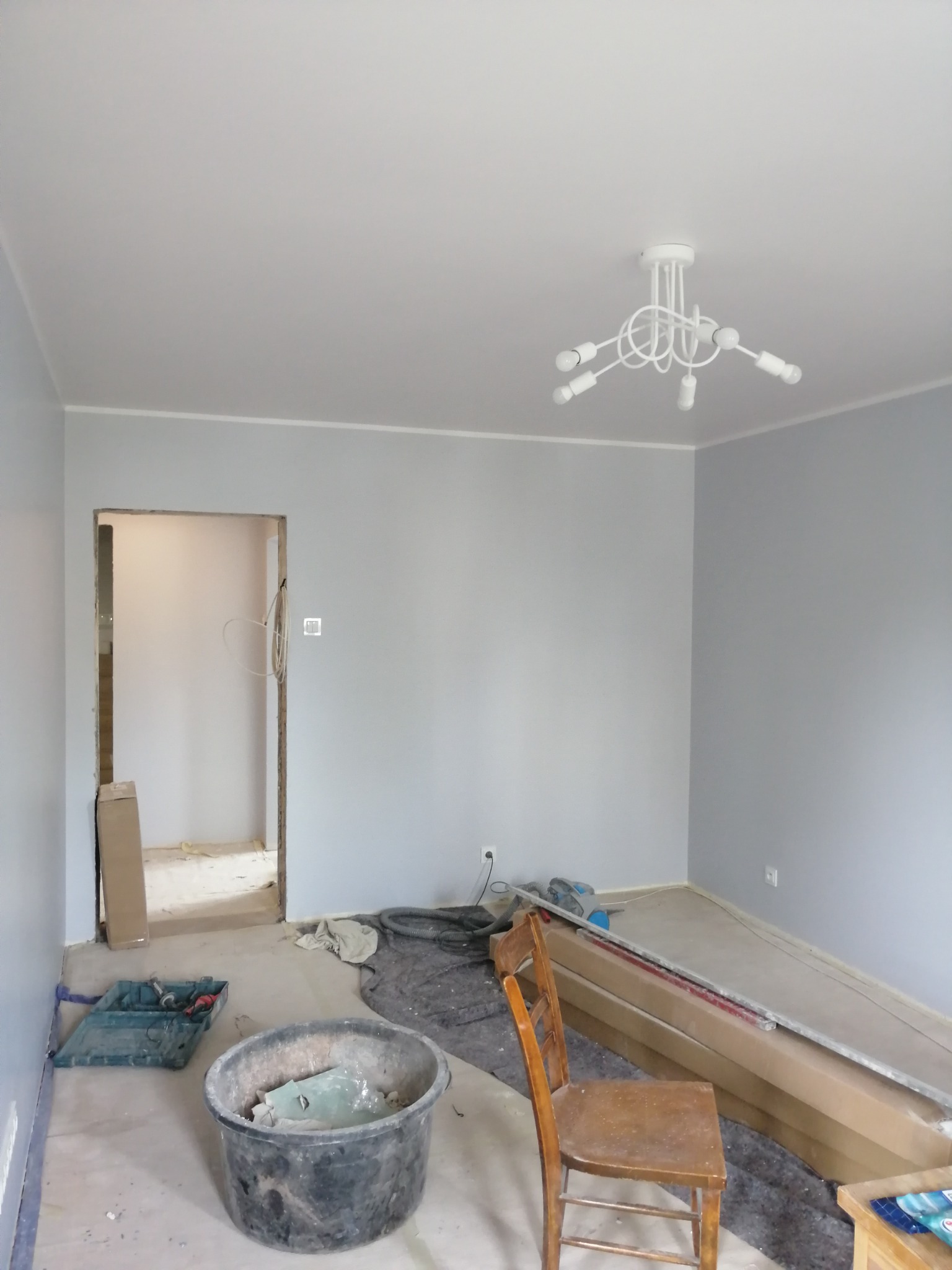 Pokój w trakcie prac wykończeniowych: szare ściany, biała lampa sufitowa, wiadro z odpadami budowlanymi, kartony z materiałami na podłodze, krzesło i widoczne ślady remontu.