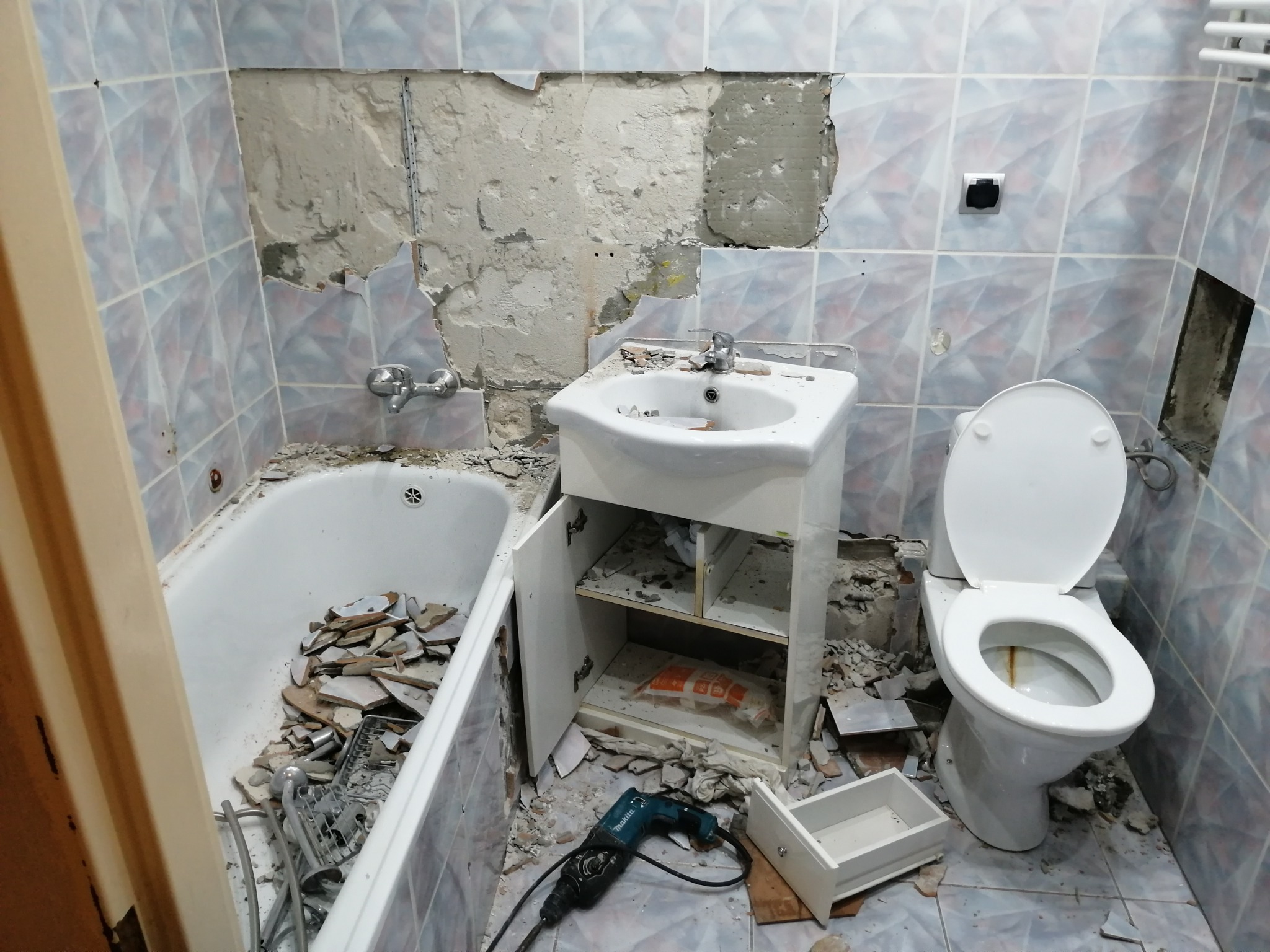 Demontaż płytek w łazience podczas remontu, widoczna wanna wypełniona gruzem, zdemontowana umywalka z szafką, toaleta i młot udarowy Makita na podłodze.