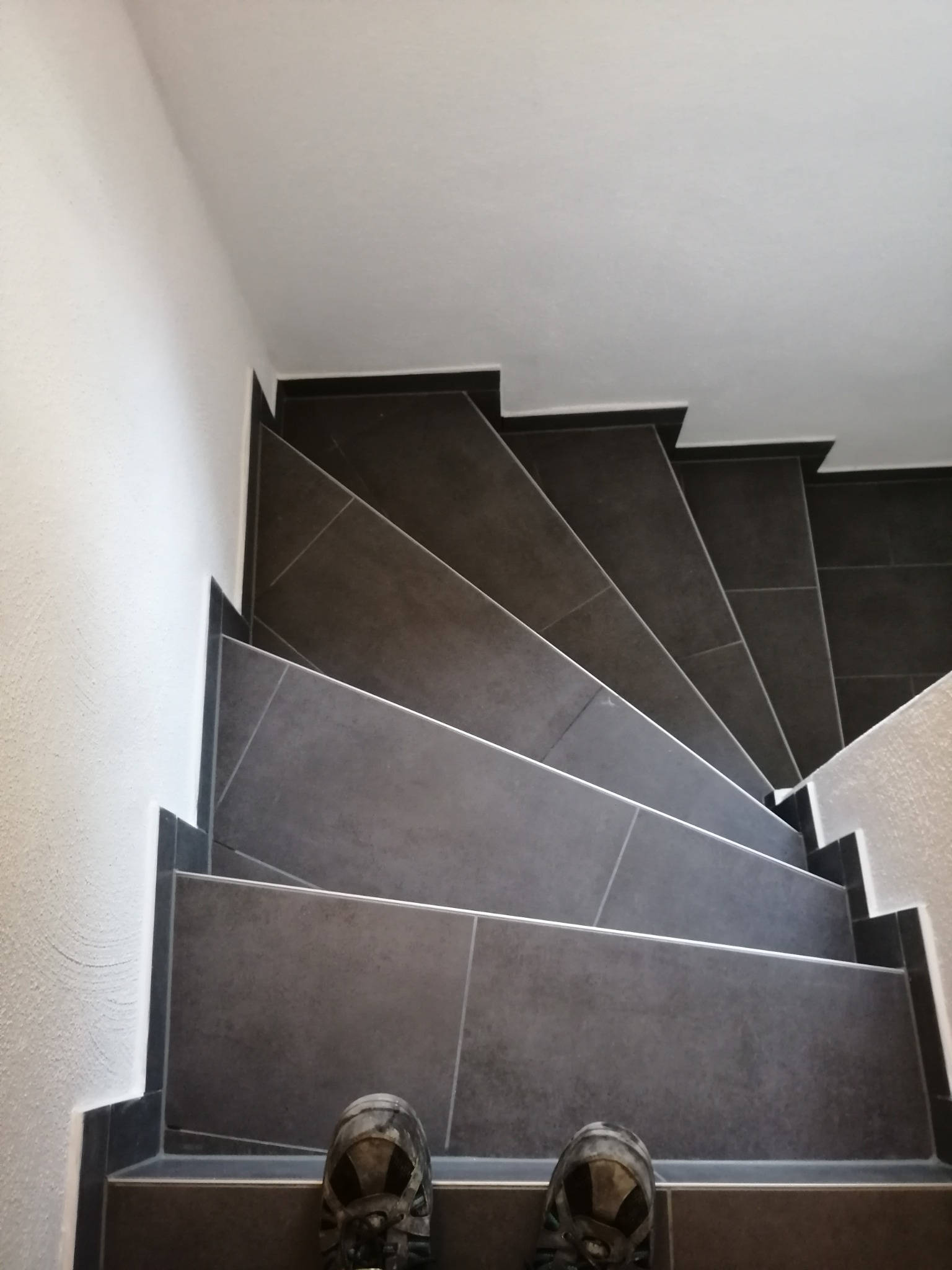 Klatka schodowa wykończona ciemnoszarą, matową glazurą, widok z góry na stopnie i fragment butów osoby stojącej na schodach.