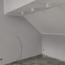 Pomieszczenie na poddaszu po remoncie, świeżo pomalowane na szaro, z zamontowanymi lampami punktowymi w suficie, widoczne wystające kable i szare płytki na podłodze.