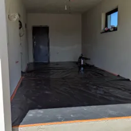 Przygotowanie podłoża w pomieszczeniu: rozłożona czarna folia ochronna z pomarańczową taśmą na obwodzie, szlifierka do gładzi na środku, surowe ściany i schody wejściowe.