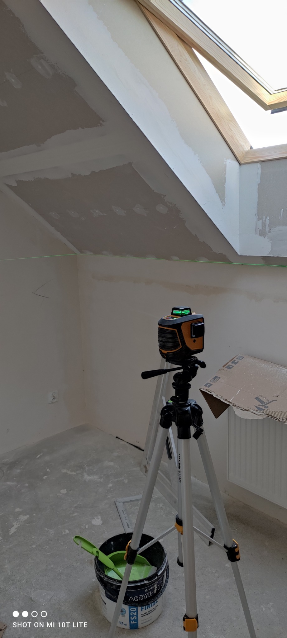 Poddasze w trakcie prac wykończeniowych, widoczny laser krzyżowy na statywie, wiadro z zieloną farbą i szpachelką, sufit z oknem dachowym w trakcie szpachlowania, ściany przygotowane do malowania.