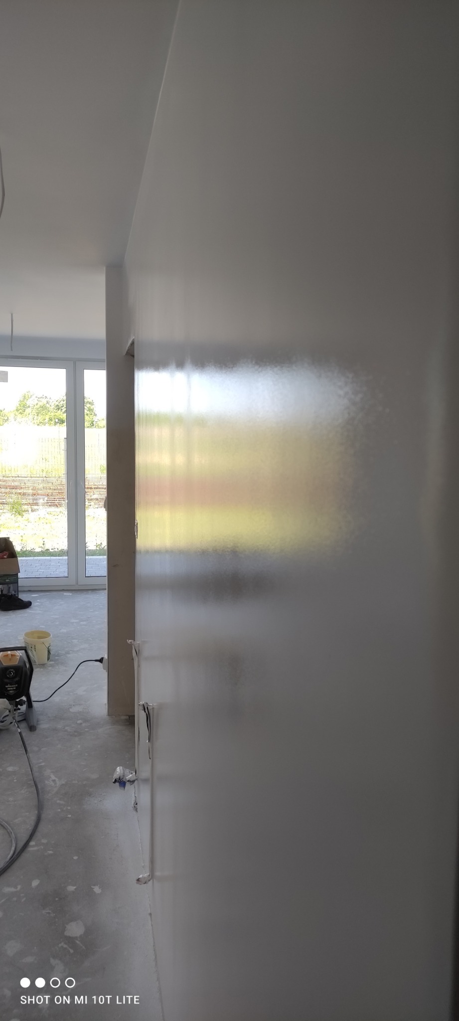 Świeżo pomalowana ściana w jasnym odcieniu, widoczny sprzęt do malowania natryskowego i wiadro z farbą, okno z widokiem na zieleń w tle.