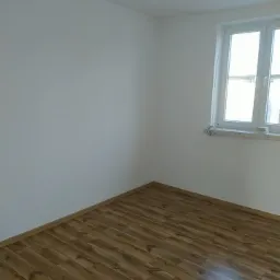 Pusty, odnowiony pokój z drewnianą podłogą z paneli i świeżo pomalowanymi białymi ścianami, widok na okno z marmurowym parapetem.