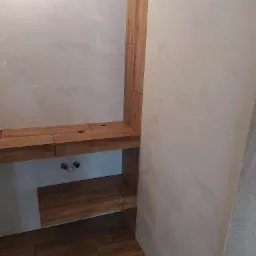 Wykończona łazienka z drewnianą zabudową wnęki prysznicowej, marmurowe płytki ścienne i podłogowe imitujące drewno, widoczne przyłącza wodne.