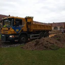 Żółto-niebieska ciężarówka Scania z wywrotką na terenie budowy, z wykopaną ziemią i widoczną koparką w tle, na tle budynku z czerwonym dachem.