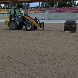 Żółta ładowarka teleskopowa Kramer Allrad 850 na piaszczystym terenie stadionu, obok paleta z rolkami trawy, trybuny w tle.