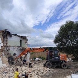 Demontix - Pomarańczowa koparka Hitachi wyburza budynek, tworząc stos gruzu z cegieł i betonu. Robotnicy ręcznie usuwają resztki, a w tle widać pole i drzewo pod zachmurzonym niebem.