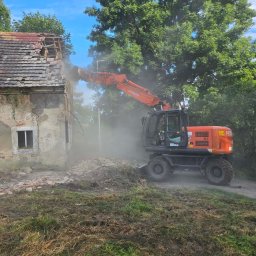 Demontix - Pomarańczowa koparka kołowa Hitachi wyburza dach starego, zniszczonego budynku, unosząc tumany kurzu. W tle zielone drzewa i błękitne niebo.
