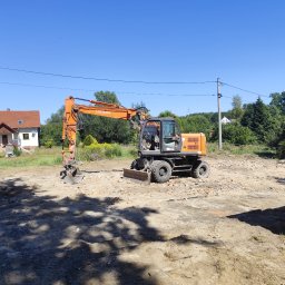 Demontix - Pomarańczowa koparka kołowa Hitachi z lemieszem spycharkowym, stojąca na placu budowy pełnym gruzu i ziemi, z domem i drzewami w tle pod błękitnym niebem.