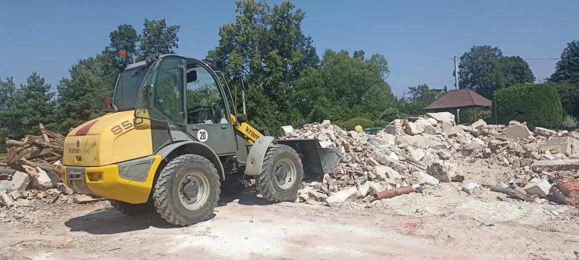 Żółto-szara ładowarka Kramer 850 na tle gruzowiska po rozbiórce, z zielenią w tle i błękitnym niebem. Widoczny fragment drewnianych pozostałości.