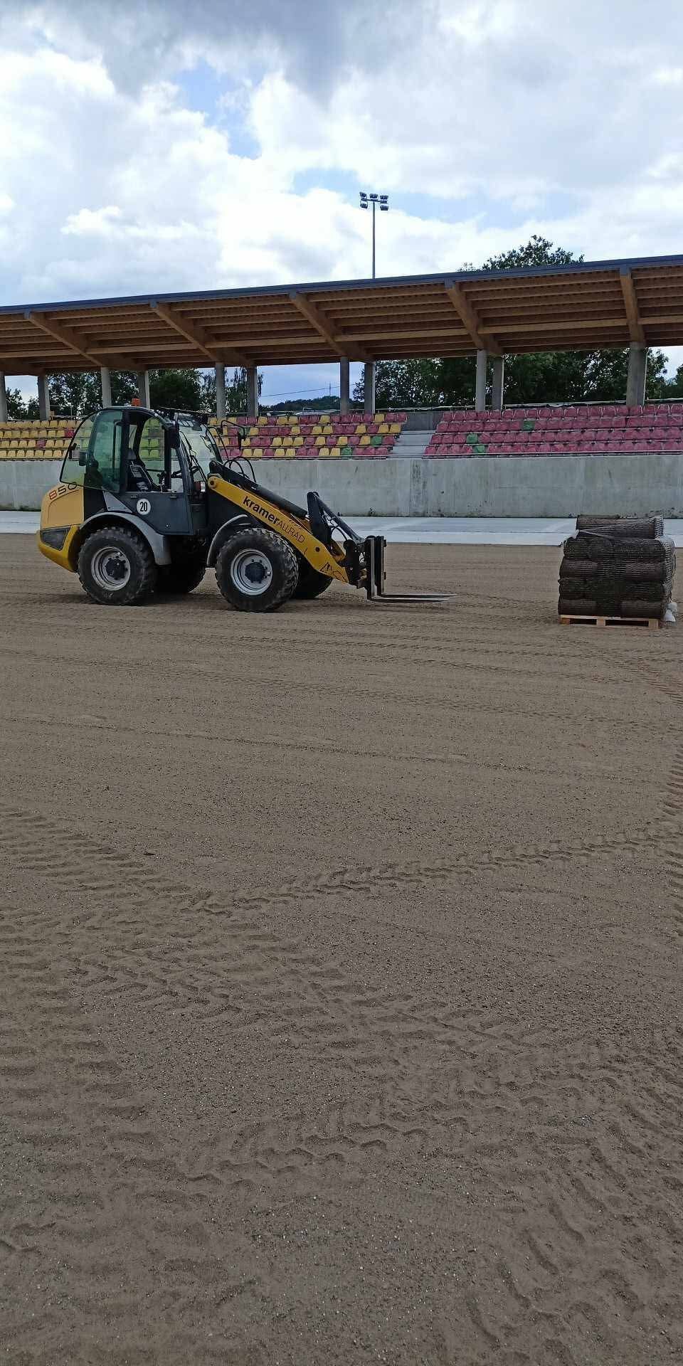 Żółta ładowarka teleskopowa Kramer Allrad 850 na piaszczystym terenie stadionu, obok paleta z rolkami trawy, trybuny w tle.