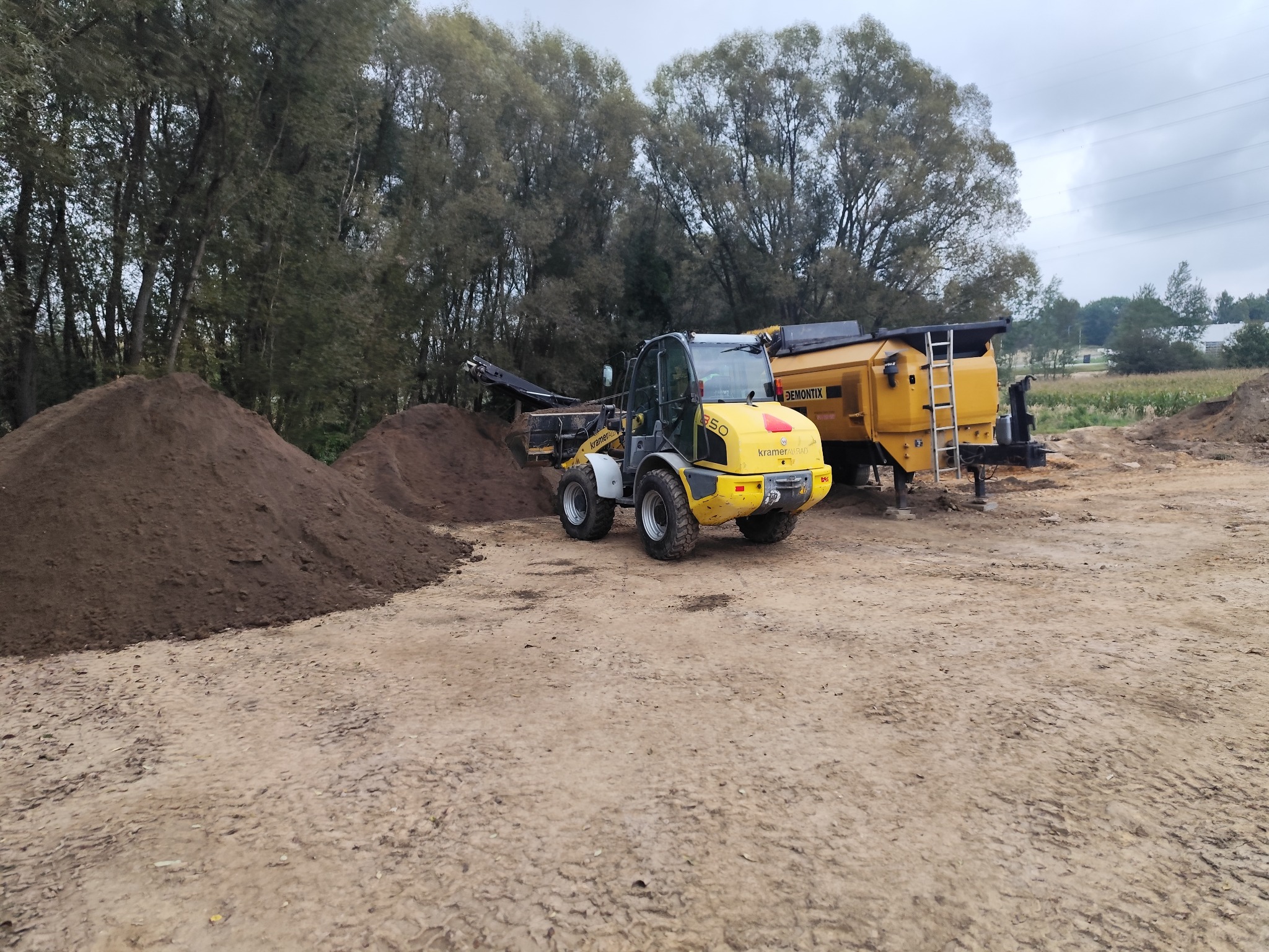 Żółta ładowarka Kramer 350 i przesiewacz do ziemi na placu budowy, obok pryzma ziemi. W tle drzewa i pochmurne niebo.