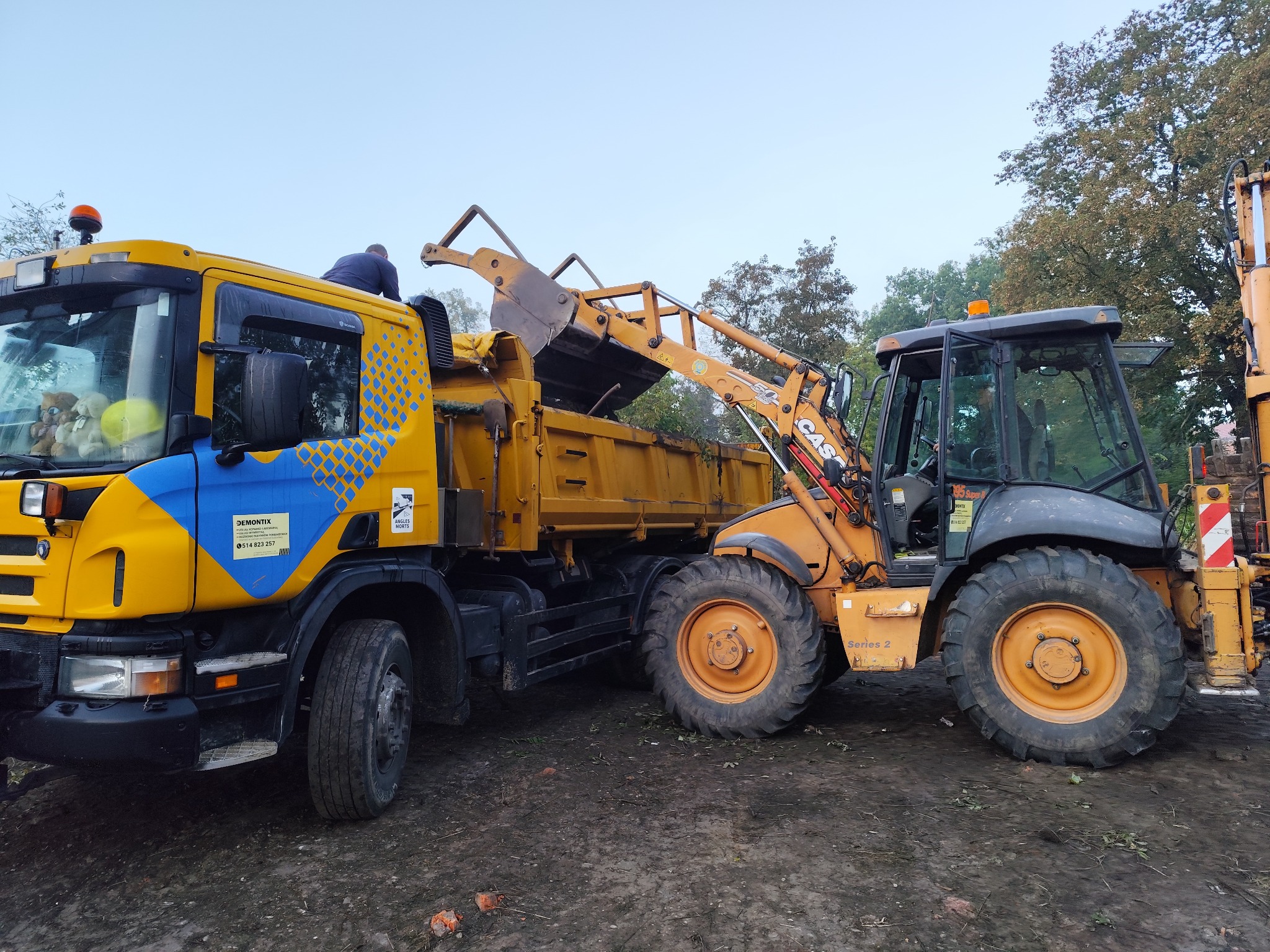 Żółty ciągnik z łyżką ładuje ziemię do ciężarówki z niebieskimi akcentami. Widoczny operator na ciężarówce i teren z resztkami cegieł.