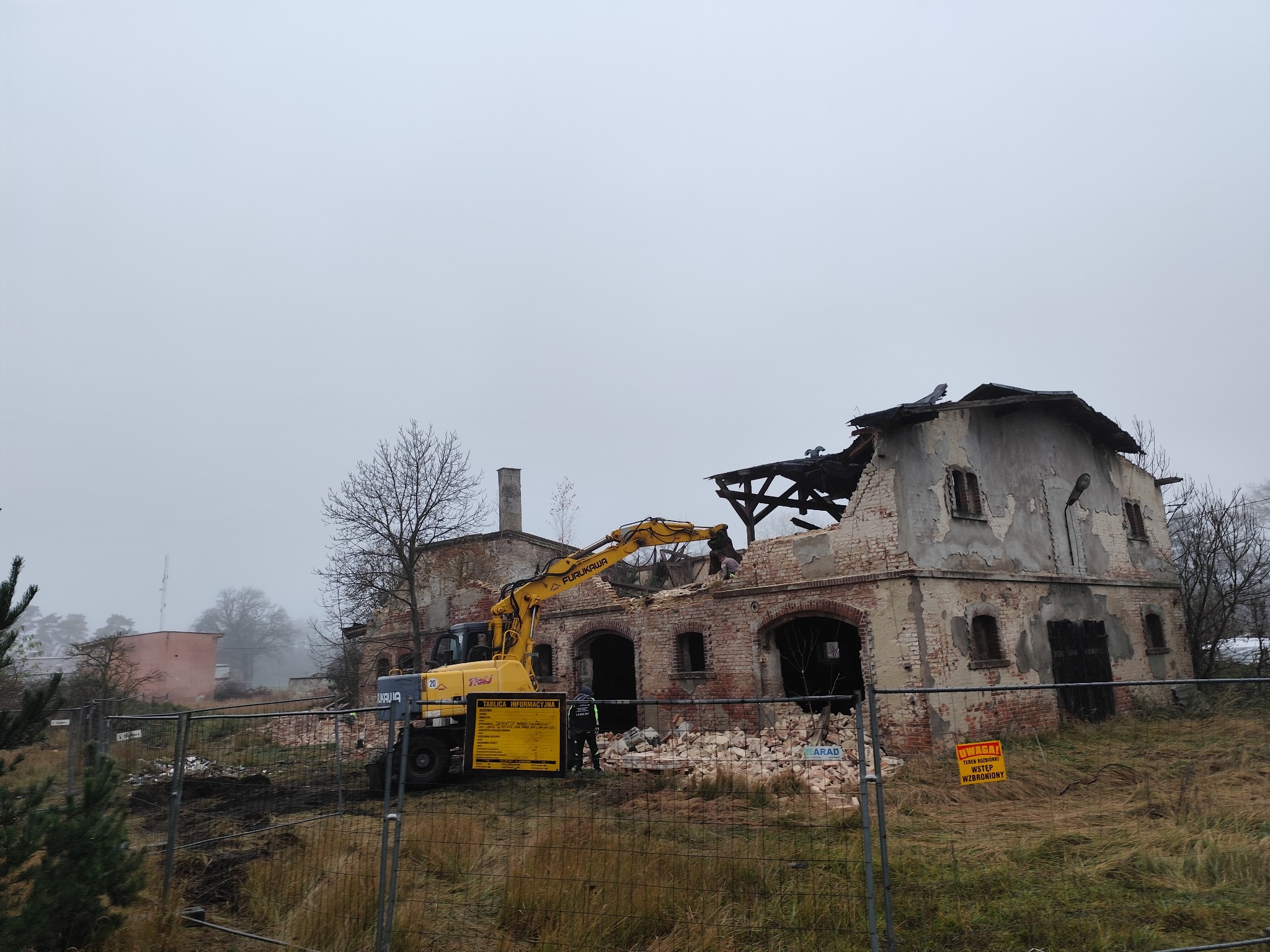 Żółta koparka Furukawa rozbiera ceglany budynek z dziurami w dachu i ścianach. Teren zabezpieczony siatką i tablicami ostrzegawczymi. Mglista pogoda.
