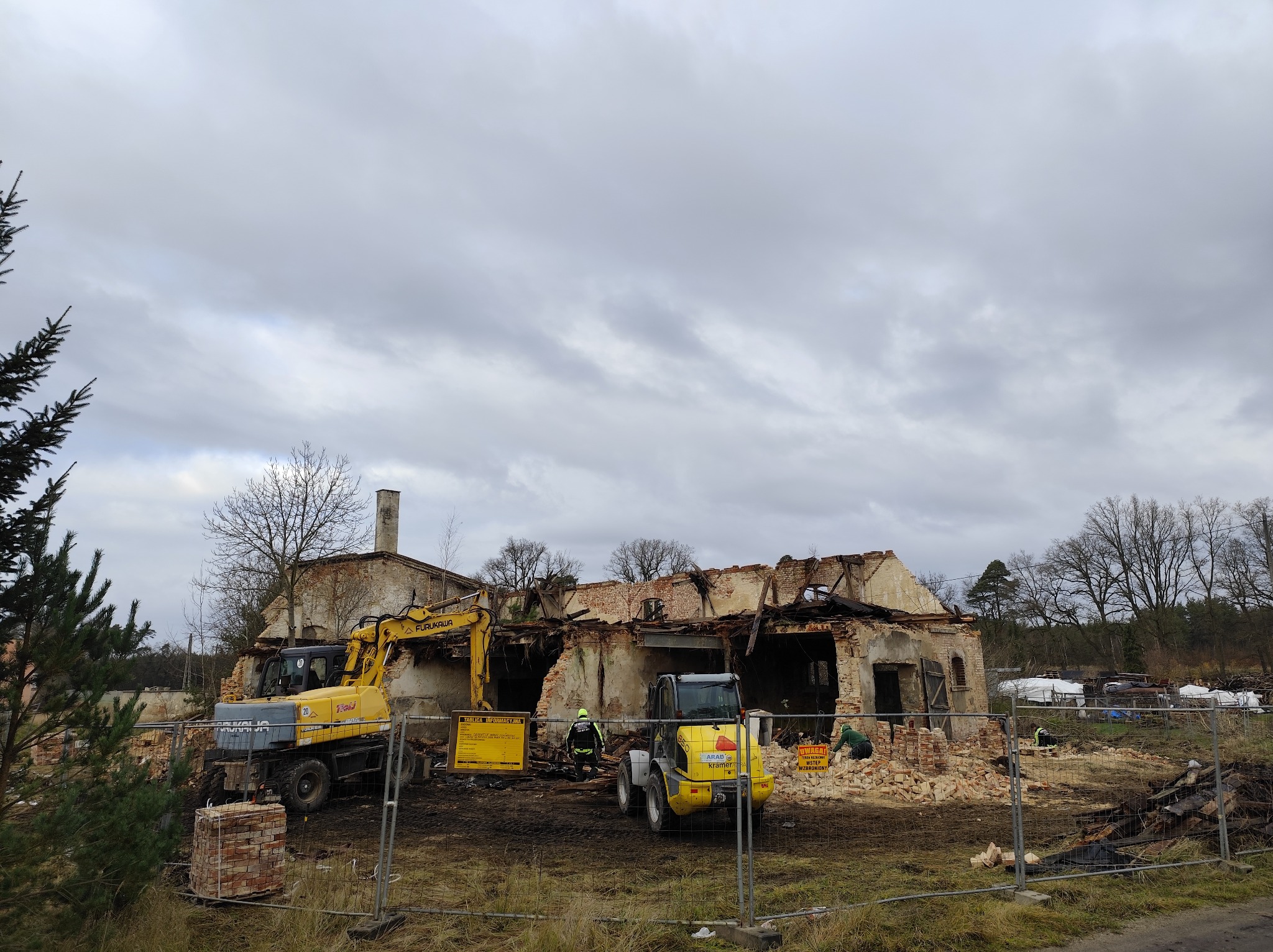Rozbiórka starego, ceglanego budynku z użyciem dwóch żółtych maszyn budowlanych. Widoczne gruzy i zabezpieczenia terenu. Pochmurne niebo w tle.