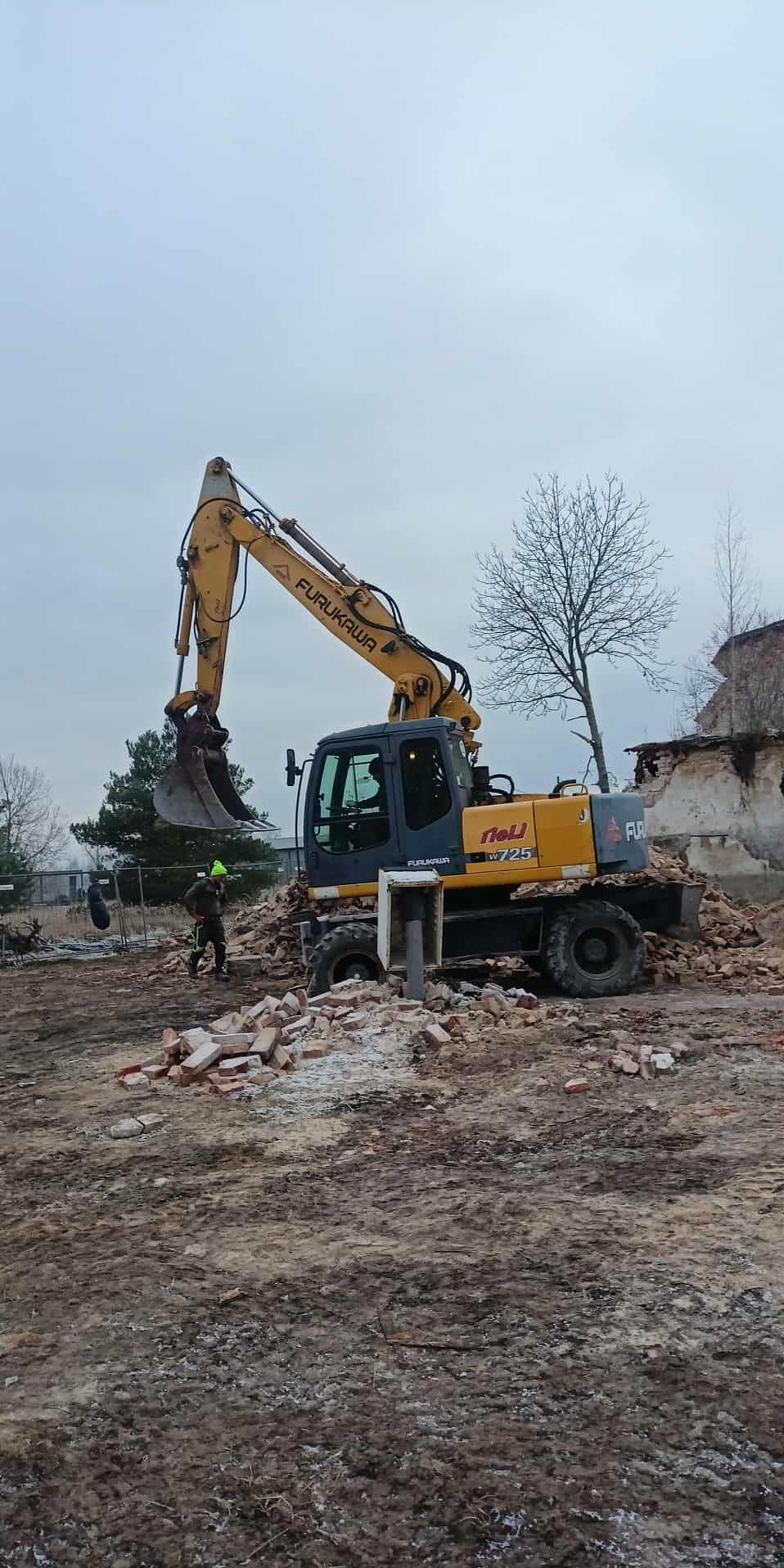 Żółta koparka Furukawa W725 w trakcie rozbiórki ceglanego budynku, z pracownikiem w zielonej czapce w tle, na placu budowy z gruzem i błotem.