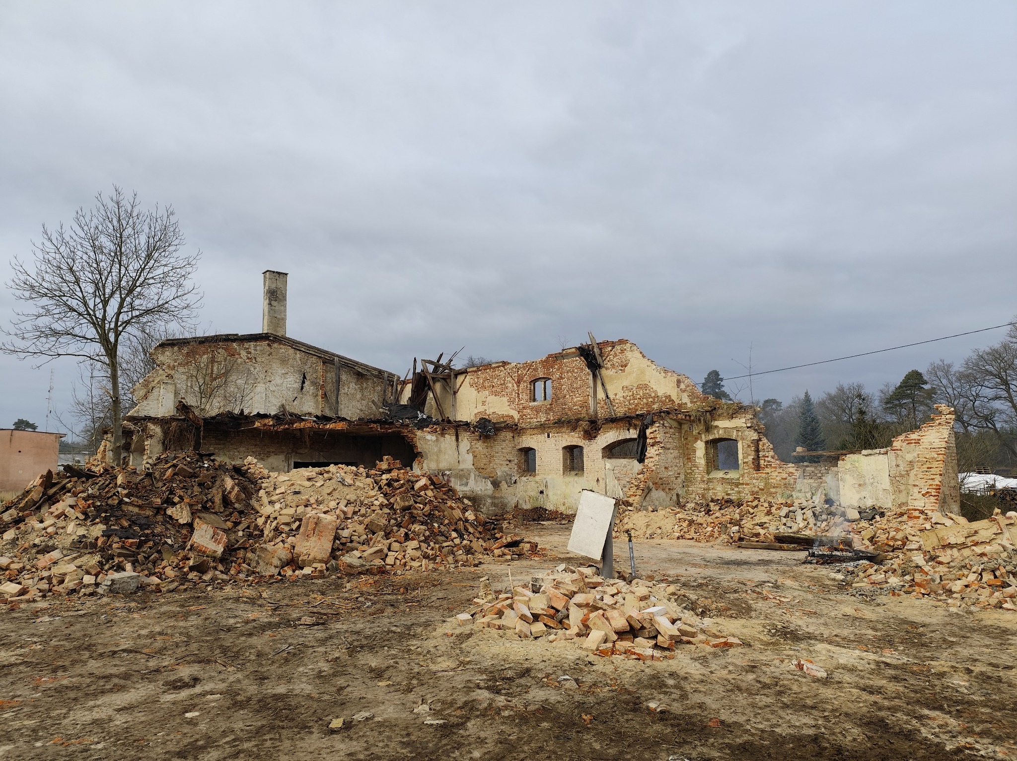 Zrujnowany budynek z cegły po rozbiórce, z widocznymi stertami gruzu i fragmentami ścian. W tle drzewa i pochmurne niebo.