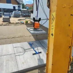Układanie szarych płyt chodnikowych z użyciem pomarańczowej zagęszczarki wibracyjnej podwieszonej na haku koparki, widoczny niebieski chwytak do kostki i zapasy kostki w tle.