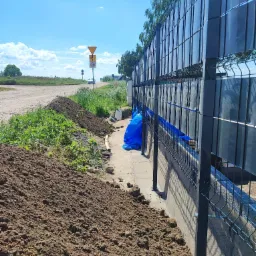 Montaż panelowego ogrodzenia z podmurówką betonową i częściowo zasłoniętymi panelami, widoczny stos ziemi i niebieska plandeka w tle, znak drogowy i droga gruntowa.