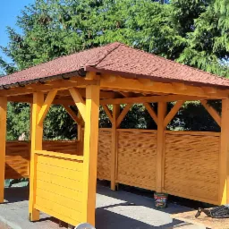 Żółta, drewniana altana z brązowym dachem, częściowo osłonięta, z widokiem na jezioro i samochód w tle. Widoczne narzędzia i farba wskazują na trwające prace wykończeniowe.