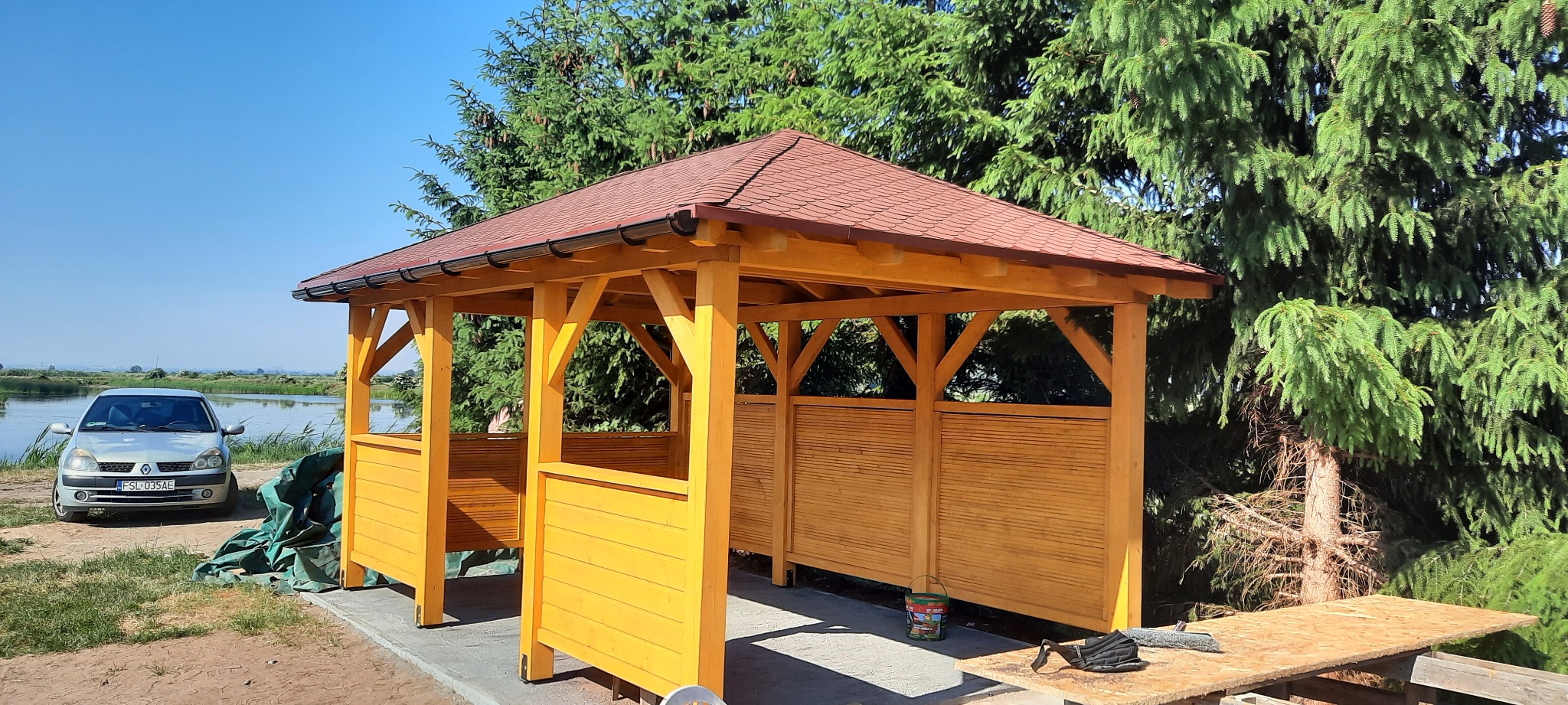 Żółta, drewniana altana z brązowym dachem, częściowo osłonięta, z widokiem na jezioro i samochód w tle. Widoczne narzędzia i farba wskazują na trwające prace wykończeniowe.