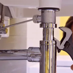 Hydraulik dokręca śrubę regulowaną czarną nasadką pod umywalką, widoczny fragment twarzy hydraulika