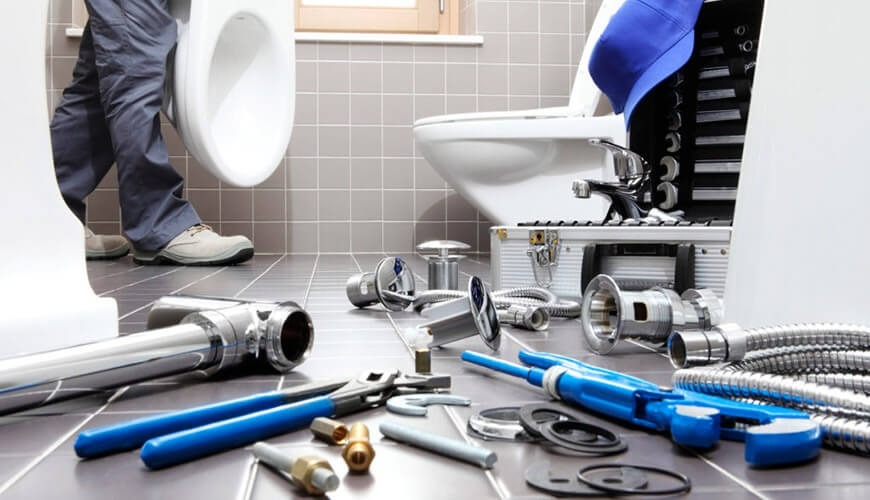 Instalacja toalety w łazience z szarymi płytkami, widoczne narzędzia hydrauliczne i akcesoria na podłodze, otwarty kufer z narzędziami w tle.