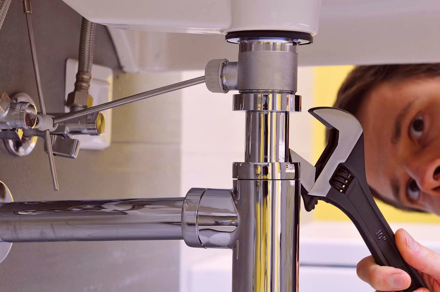 Hydraulik dokręca śrubę regulowaną czarną nasadką pod umywalką, widoczny fragment twarzy hydraulika