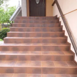 Zewnętrzne schody wejściowe do domu wyłożone brązowymi płytkami ceramicznymi, z metalową poręczą po prawej stronie i drewnianymi drzwiami wejściowymi w tle.