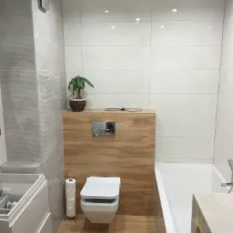 Nowoczesna łazienka z podwieszaną toaletą, drewnopodobną zabudową, białymi płytkami i falistą ścianą, uzupełniona o roślinę doniczkową i stojak na papier toaletowy.