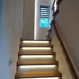 Drewniane schody z oświetleniem LED pod stopniami, prowadzące w górę, z widocznym fragmentem okna w białej ceglanej ścianie w tle.