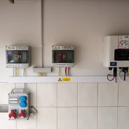 Rozdzielnice PV-AC i PV-DC, inwerter fotowoltaiczny oraz gniazda siłowe zamontowane na kafelkowanej ścianie, widoczna tabliczka ostrzegawcza o napięciu.