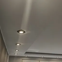 Równomiernie rozmieszczone, kwadratowe oprawy oświetleniowe w suficie podwieszanym, z widocznym światłem padającym w dół, okno z widokiem na zieleń w tle.