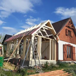 Konstrukcja drewniana dobudowywana do istniejącego domu z brązową stolarką okienną, widoczny pracownik w niebieskim kombinezonie i drabina aluminiowa.