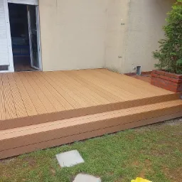 Nowoczesny taras kompozytowy z dwoma stopniami, prowadzący do drzwi balkonowych, otoczony zielenią i ceglaną donicą z tujami.