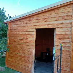 Drewniana konstrukcja z widocznym wnętrzem, prawdopodobnie w trakcie budowy, z elementami rusztowania i narzędziami w tle, otoczona zielenią.