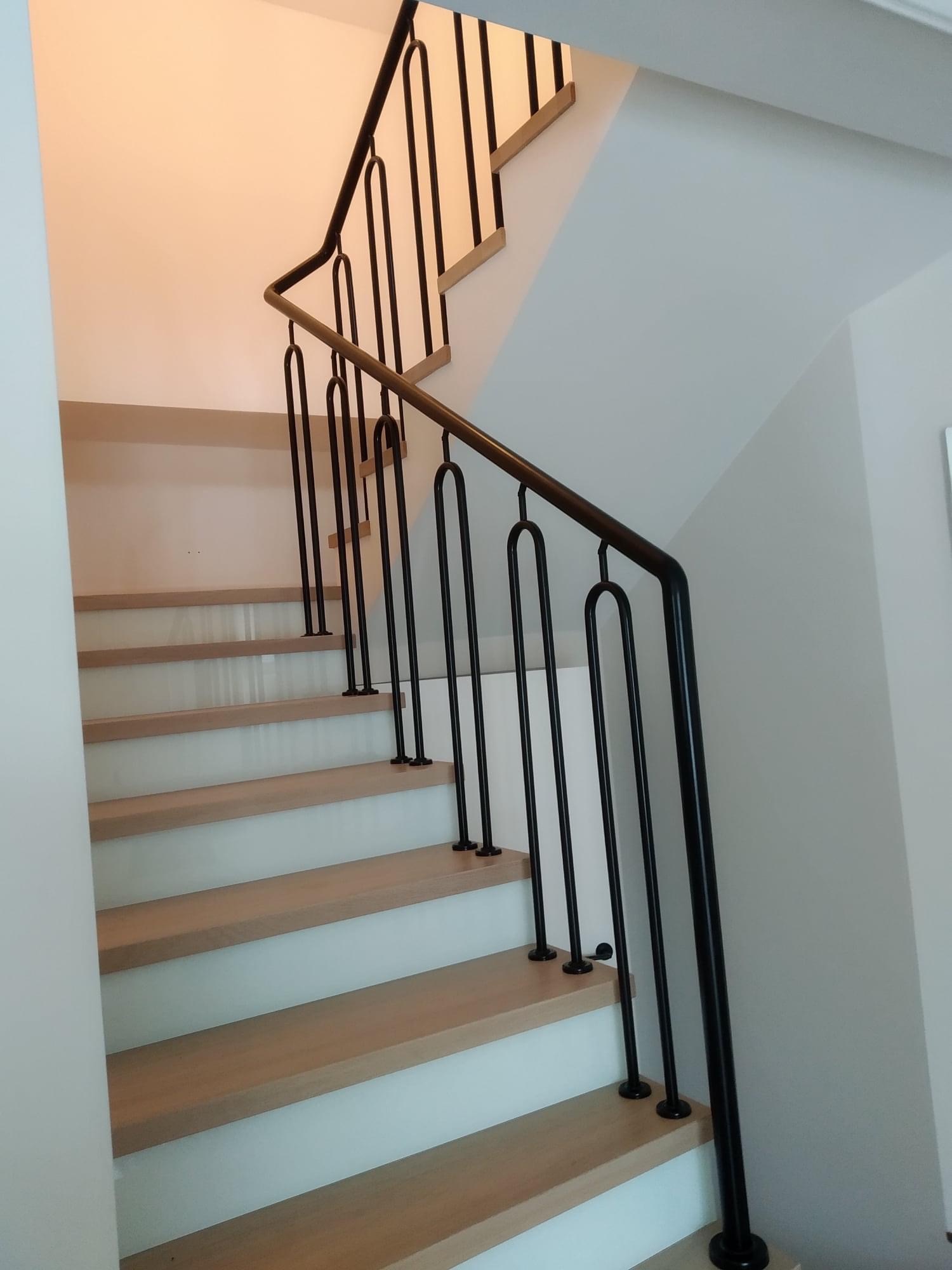 Nowoczesne schody z drewnianymi stopniami i czarną metalową balustradą z charakterystycznymi, zaokrąglonymi pionowymi elementami.