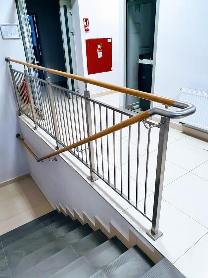 Nierdzewna balustrada schodowa z drewnianą poręczą w budynku użyteczności publicznej, widoczna gaśnica i skrzynka hydrantowa w tle.